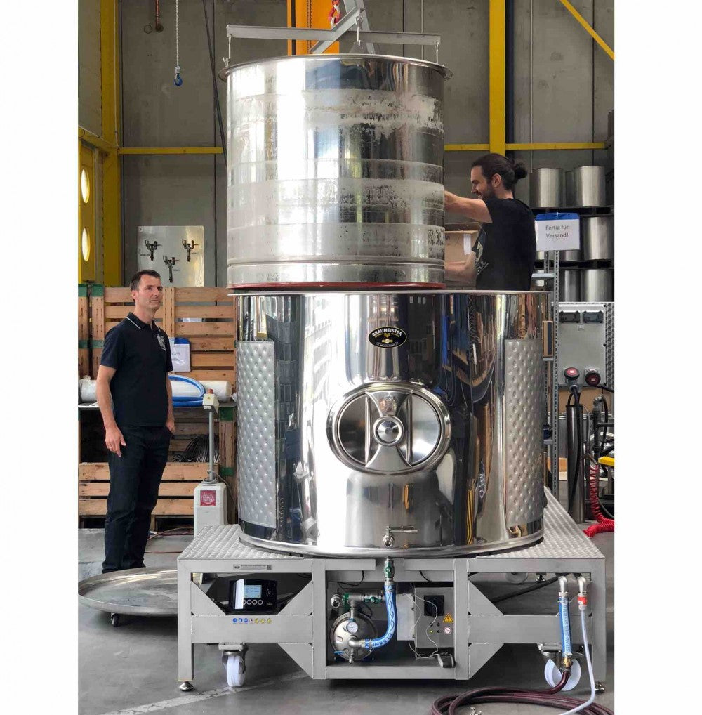 To personer betjener et Speidel #Braumeister 1000L - Bestillingsvare automatisk bryggesystem fra Speidel i et industrianlegg med gule bjelker og trepaller.