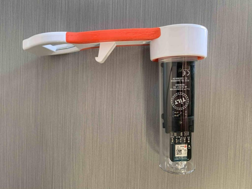 En Tilt Wrench fra Tilt Hydrometer, med et hvitt og oransje håndtak og festet til en sylindrisk, svart og gjennomsiktig del med en innebygd flaskeåpner, er montert på en grå flate.