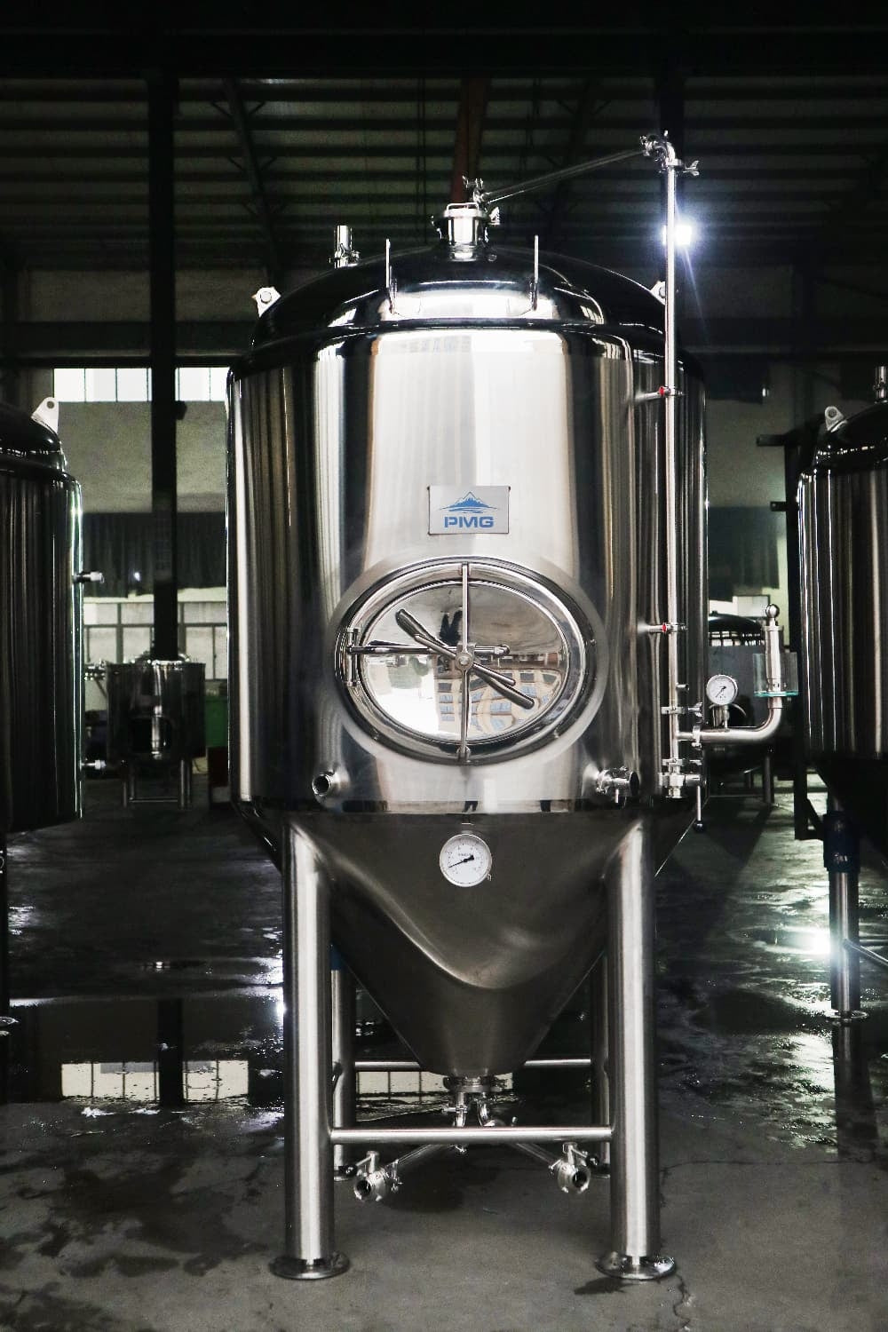 PMG 5000L Unitank av rustfritt stål, isolert og fullt utstyrt (Bestillingsvare) fra PMG, med glassluke, trykkmåler og rør, står fremtredende i et industrielt bryggeri.