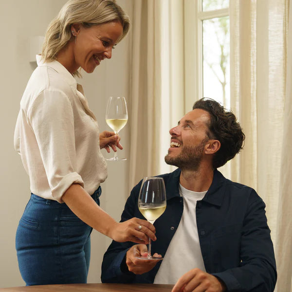 En kvinne holder et Stölzle Lausitz Fino hvitvin vinglass 450 ml og smiler til en sittende mann med et annet glass. De sitter innendørs ved et vindu med skira gardiner.