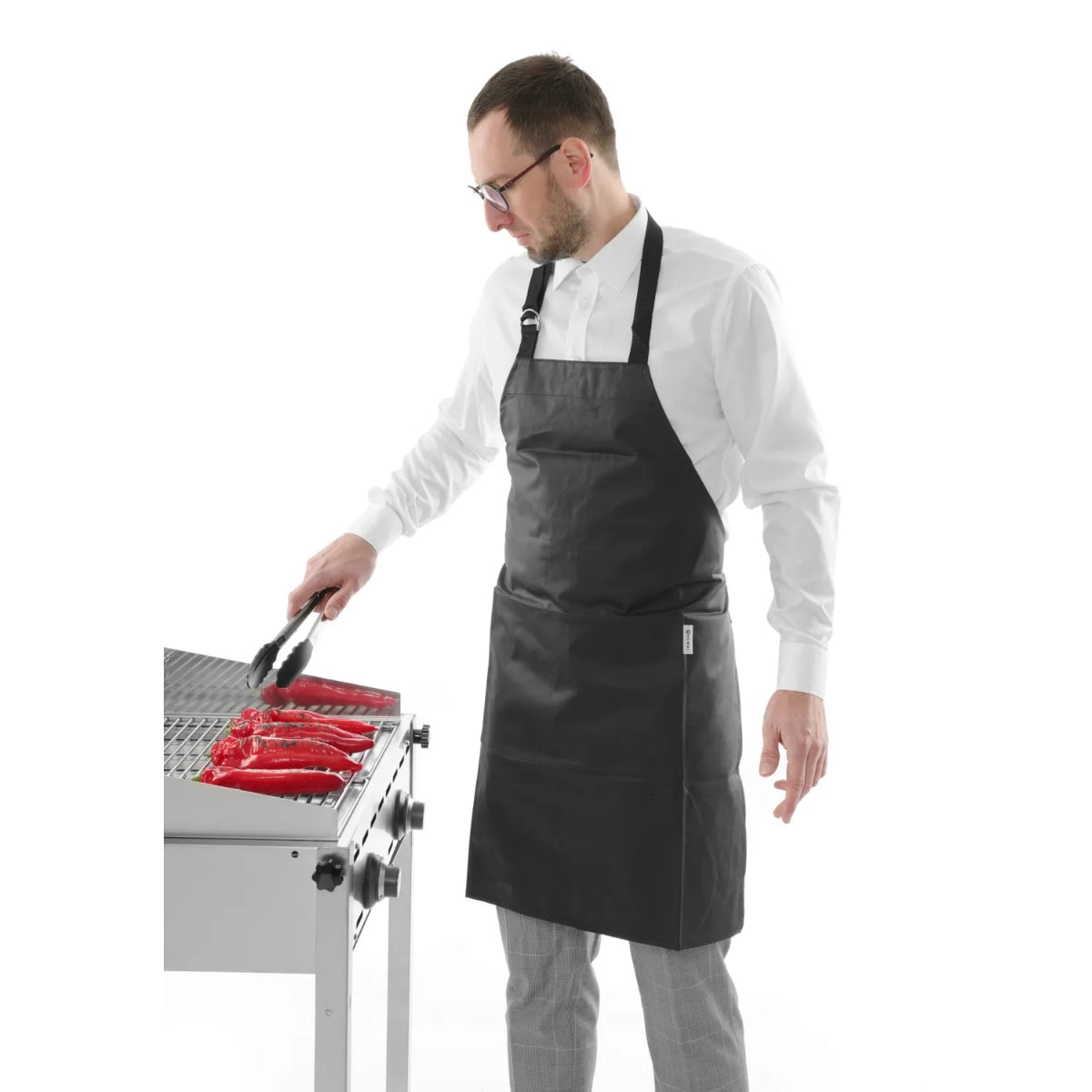En mann iført Hendi BBQ Forkle og hvit skjorte griller pølser med en tang på en grill i rustfritt stål.
