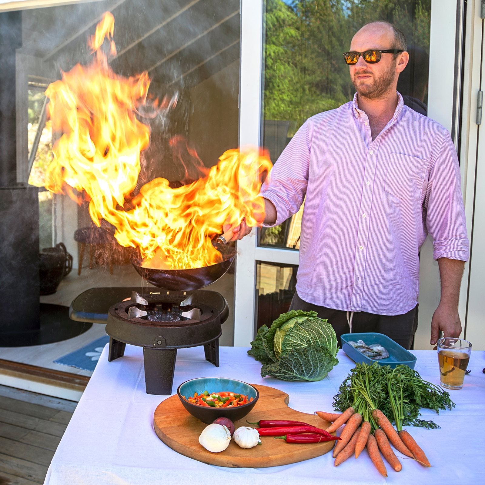 En mann med solbriller rører i en panne med grønnsaker og en drink på HOT WOK PRO 12 kW gassbrenner med wokpanne fra HotWok, og store flammer står opp mens han lager mat ved siden av bordet.
