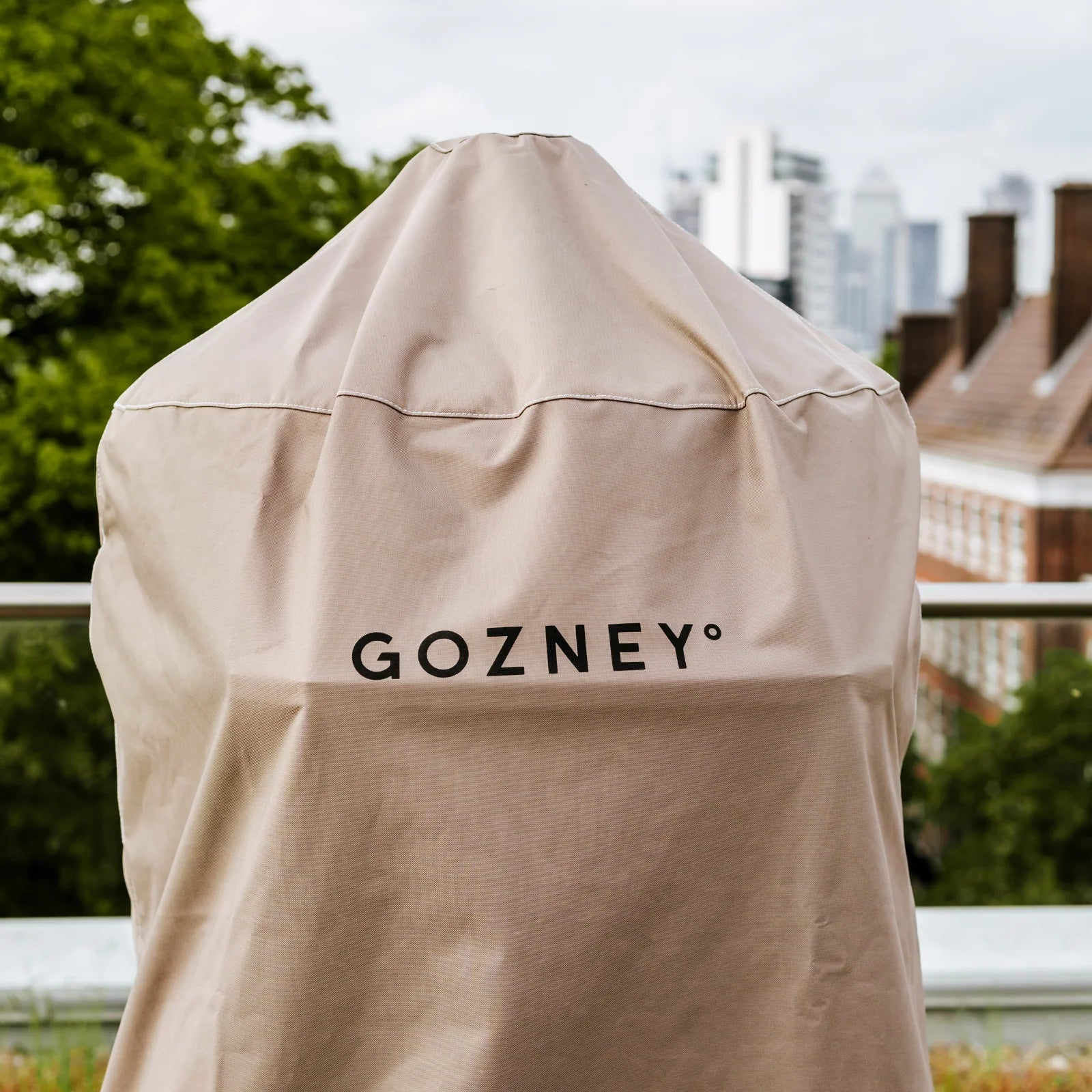 Et beige Gozney Dome & Stand Cover (Gen 1) beskytter et utendørs apparat på en balkong, med trær og bybygninger synlige i bakgrunnen.