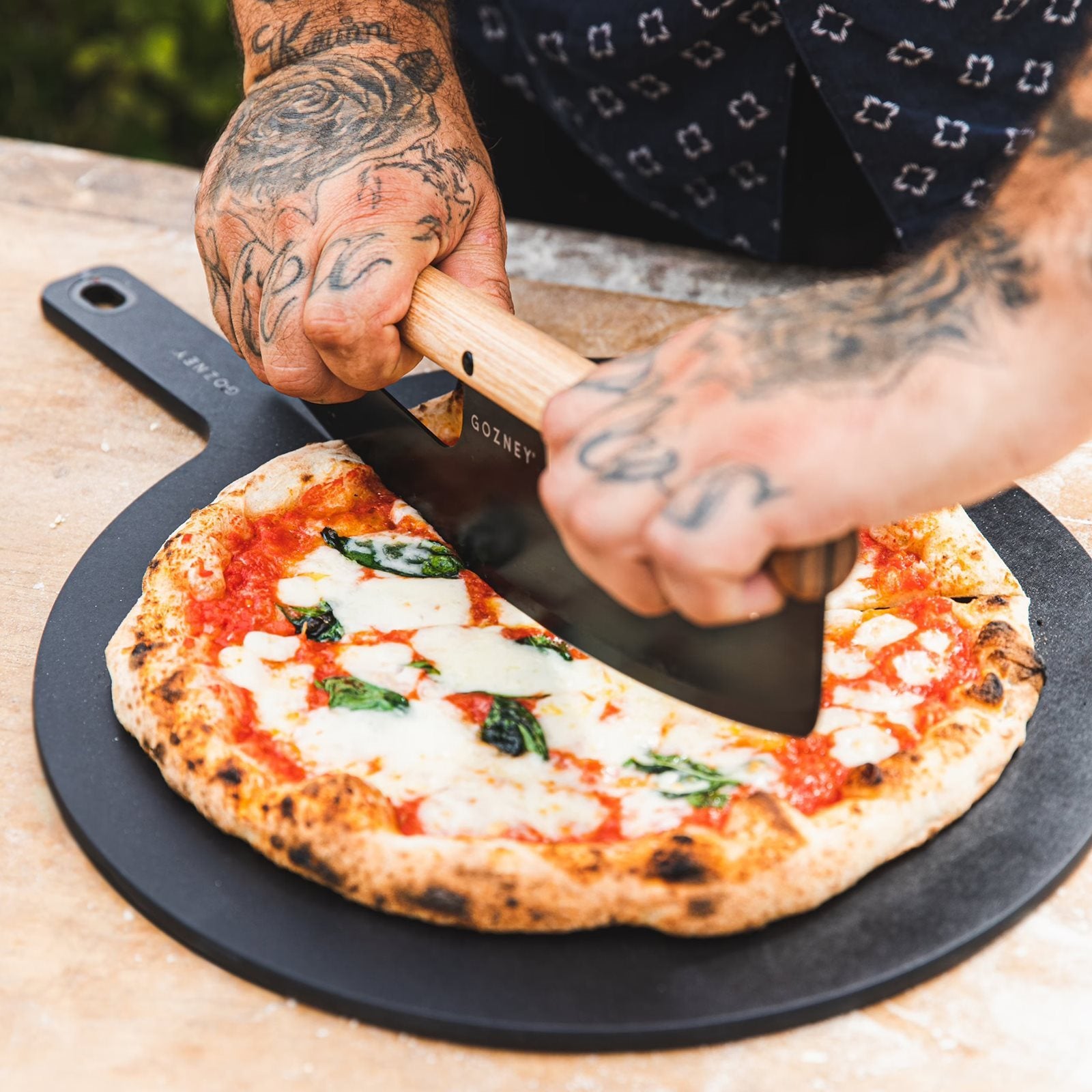 En tatovert person skjærer en Margherita-pizza med en Gozney Pizzakutter i rustfritt stål på et svart serveringsbrett.