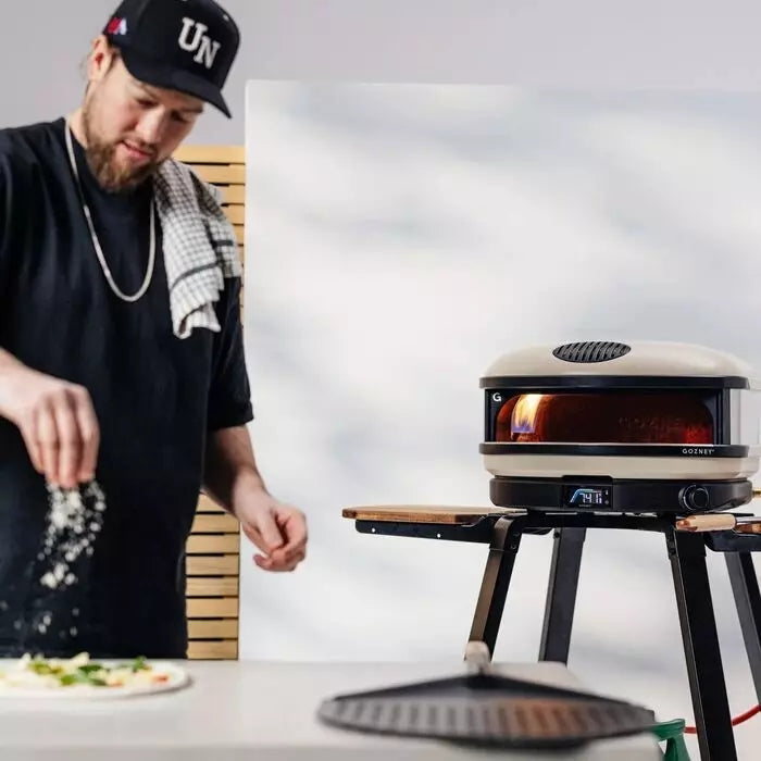 En mann drysser ingredienser på en napolitansk pizza ved siden av en Gozney Arc XL - Bone - 16" Gass Pizzaovn fra Gozney plassert på et stativ utendørs.