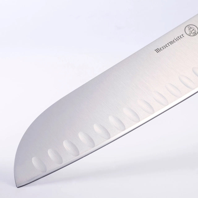 Nærbilde av bladet i rustfritt stål på Messermeister Meridian Elite 18 cm Kullenschliff Santoku japansk kokkekniv, med ovale fordypninger langs eggen og Messermeister Meridian Elite-logoen nær håndtaket.