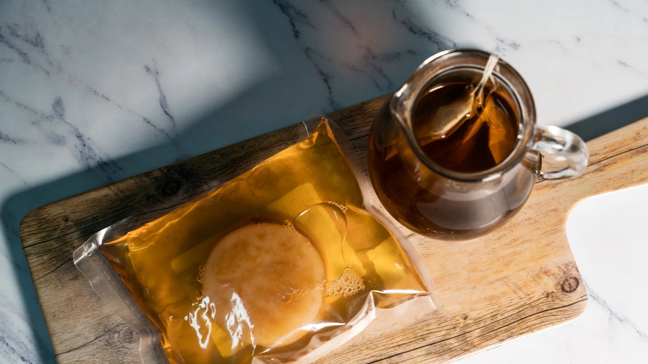 En forseglet pose med Enolandias Scoby - kultur for Kombucha (diameter 12 cm) og startvæske plasseres ved siden av et glass med te på et skjærebrett av tre, klar til fermentering.
