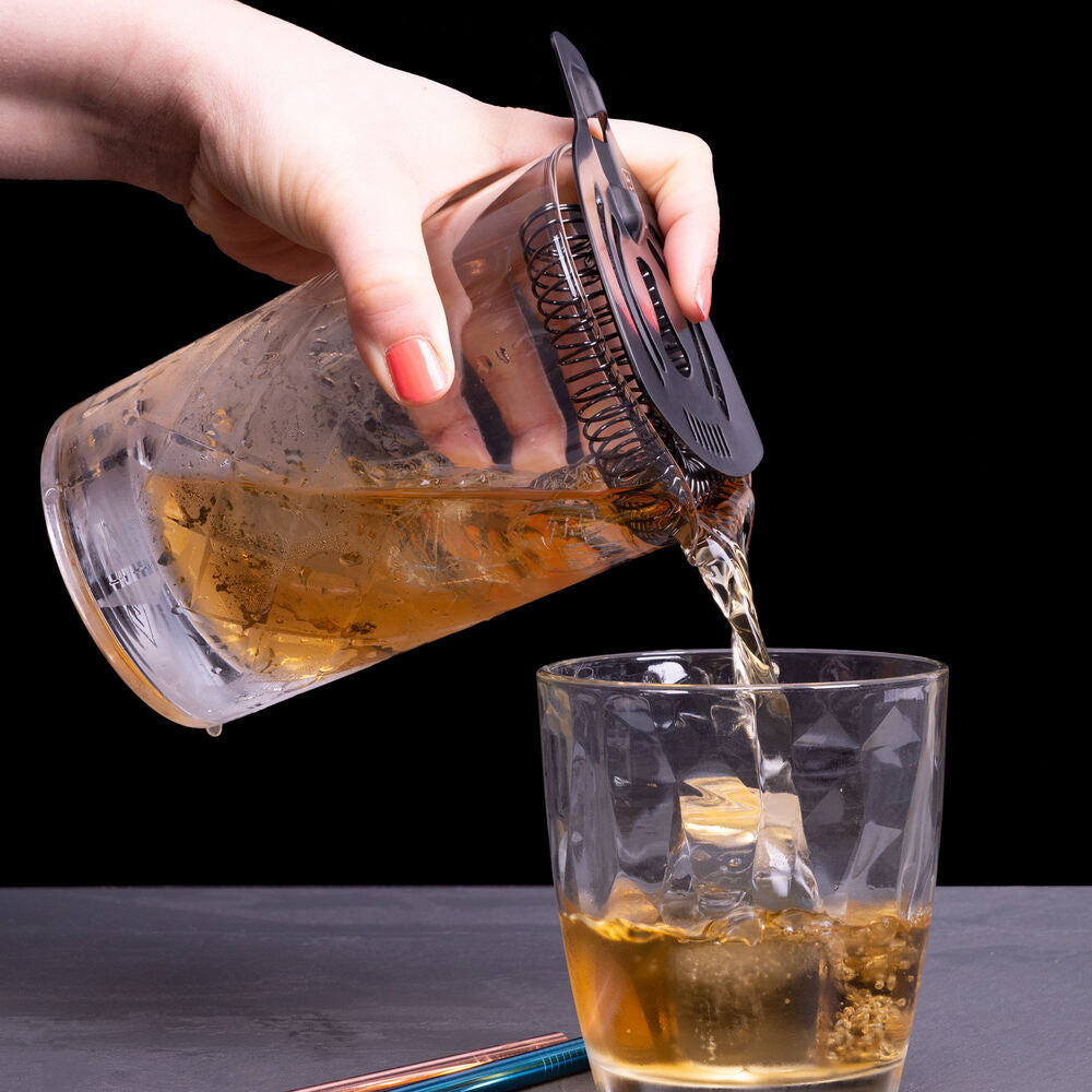 En hånd heller en gylden drink fra en cocktailshaker med sil ned i et Lurch Blandeglass på 650 ml, fylt med is, mot en svart bakgrunn – perfekt for å fremheve elegante rørte cocktailer.