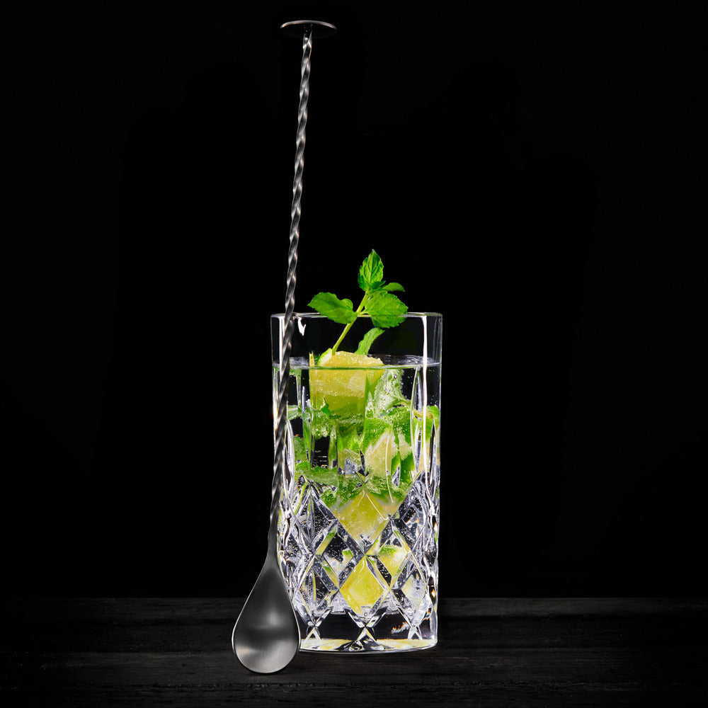 Et krystallglass fylt med en cocktail, isbiter, lime-båter og mynte, med Lurch Barskje med Muddler 29 cm plassert i glasset, mot en svart bakgrunn – det perfekte motivet for enhver cocktailbar.
