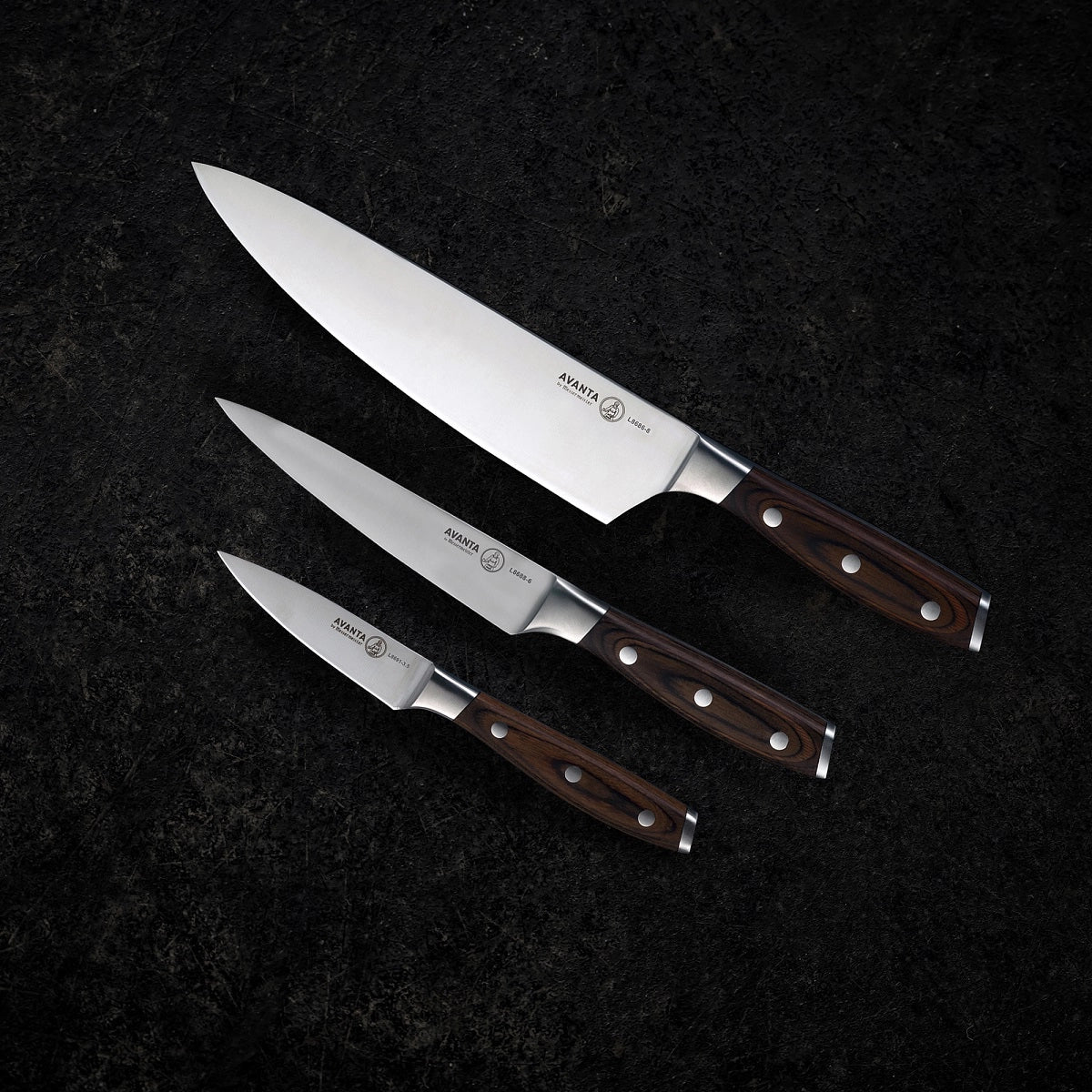 Tre Messermeister Avanta Pakkawood Knivsett-kniver med blad i tysk X50 rustfritt stål og håndtak i mørkt pakkawood er plassert parallelt på en mørk, strukturert overflate.