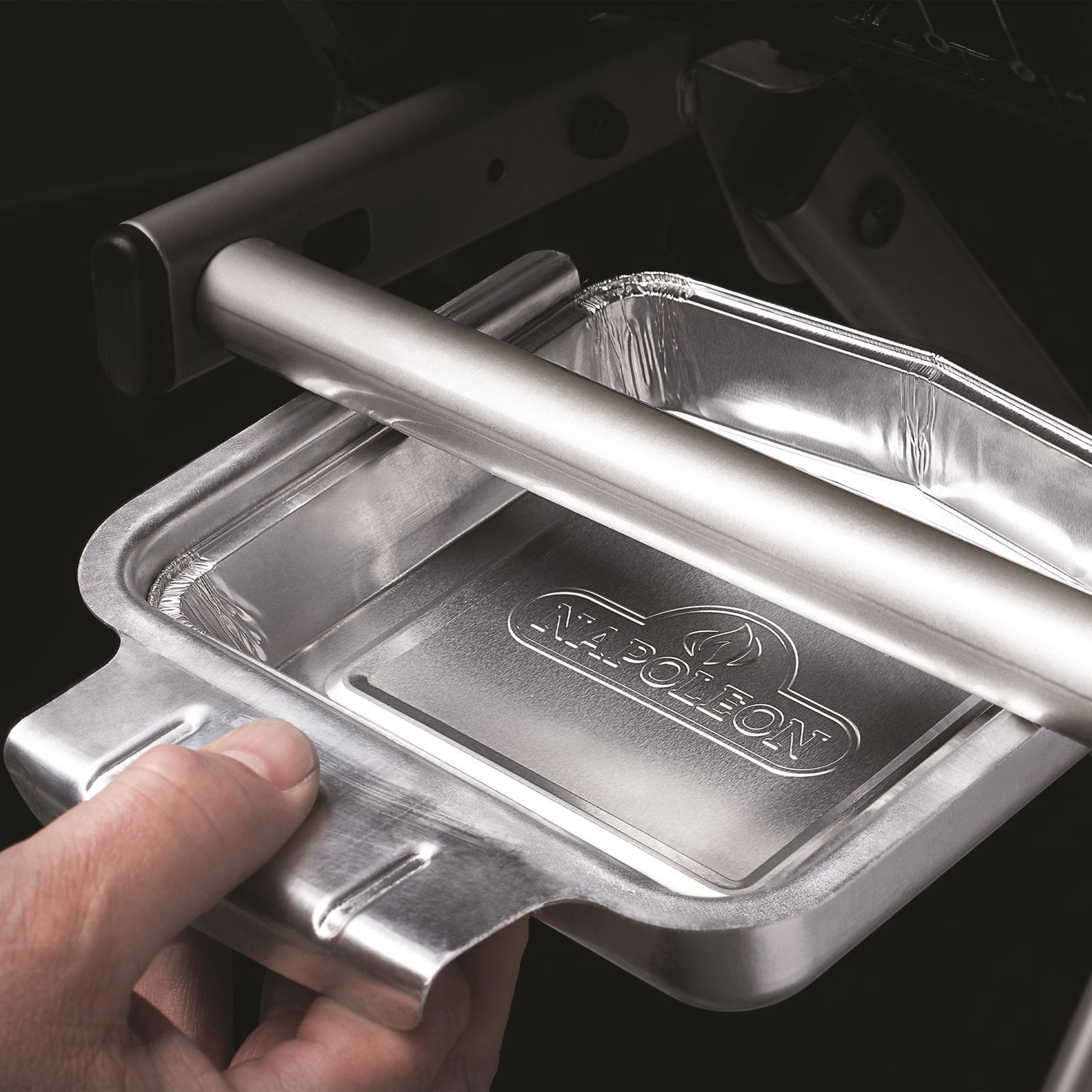 En hånd plasserer en Napoleon Aluminiumsformer TravelQ 285 aluminiumsform under en grill, ideell som TravelQ 285 tilbehør. Pakken inneholder 5 stk.