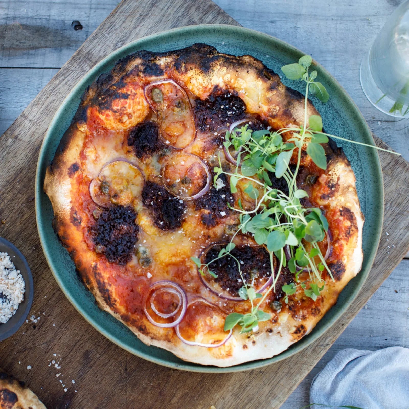 En rustikk pizza toppet med tomatsaus, ost, rødløk, friske grønnsaker og krydret Ask Nduja 100g Salami med Chili på en grønn tallerken over et underlag av tre.