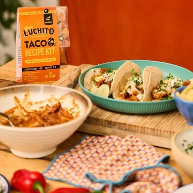 Tre ferdiglagde meksikanske tacoer på en blå tallerken, en blandeskål med fylt av marinert svinekjøtt, en eske med Gran Luchito Al Pastor Recipe Kit 300 g og et fargerikt kjøkkenhåndkle på et trebord.