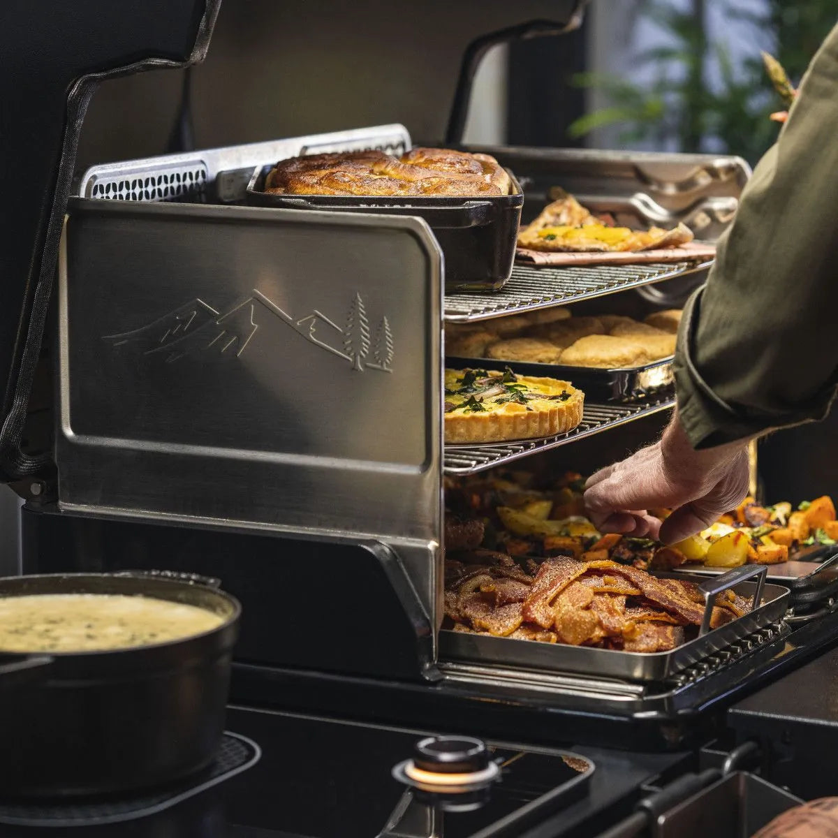 En person plasserer et brett med stekte grønnsaker i en Traeger Timberline L pelletsgrill med flere lag fylt med ulike matretter, mens en gryte putrer på komfyrtoppen ved siden av - perfekt for utendørs matlaging.