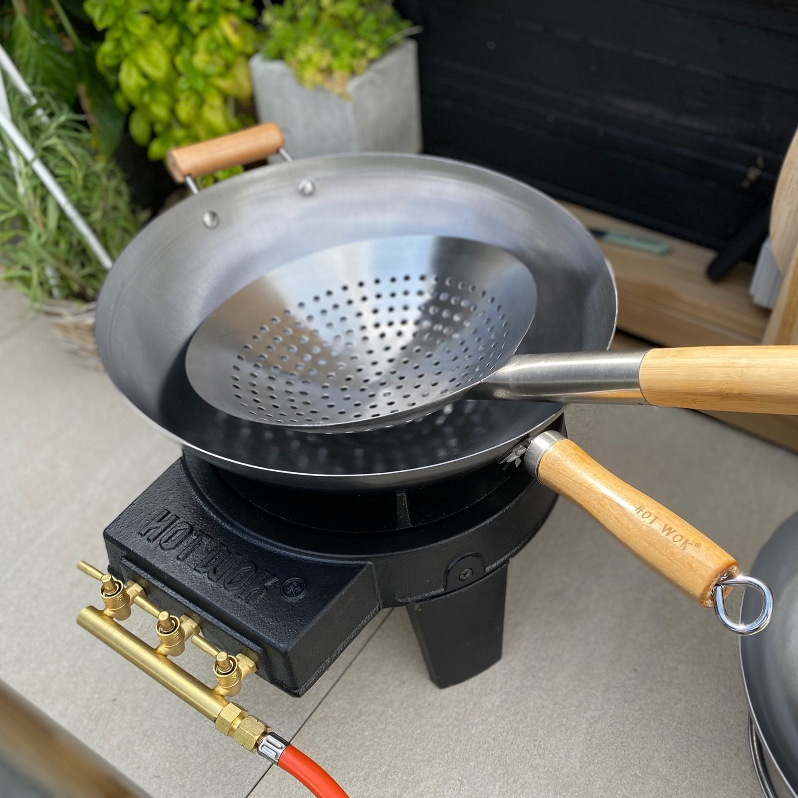 HotWok Wok Hullskje og en woksil i metall, begge med trehåndtak, står på en gassbrenner blant grønne planter og utekjøkkenutstyr.