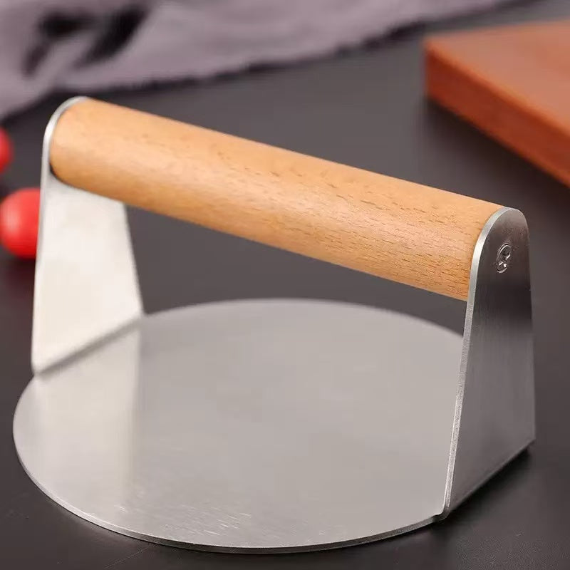 Stainless steel smash burger press with wooden handle on a dark kitchen counter