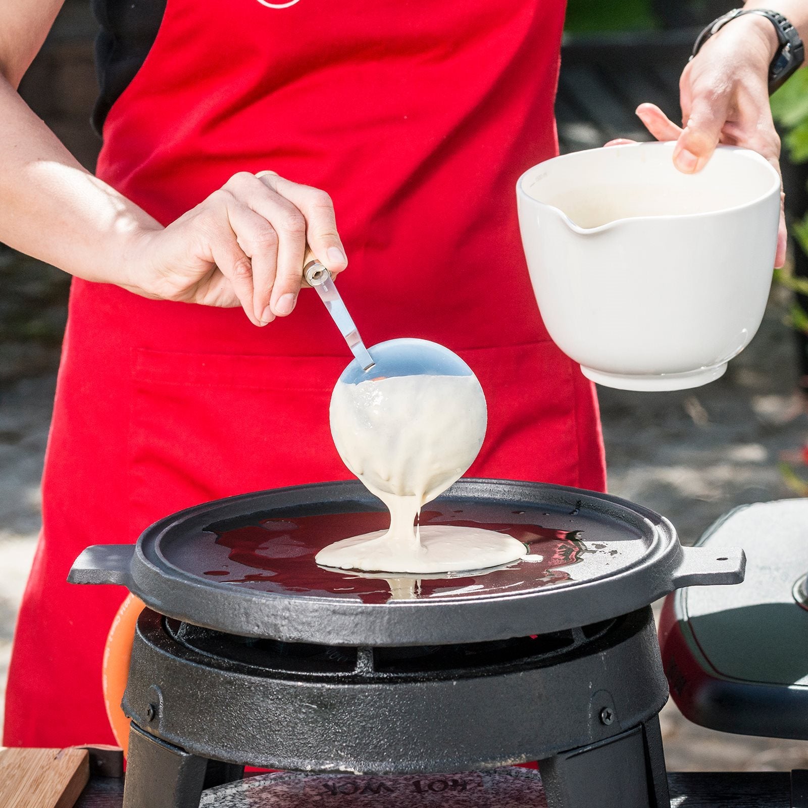 Iført rødt forkle heller en person pannekakerøre fra en øse på en Grillpanne 32,5 cm fra HotWok, plassert over HOT WOK gassbrennere utendørs.