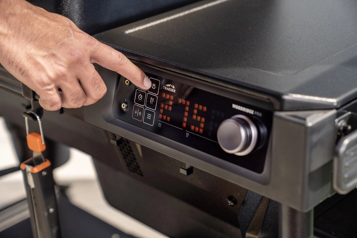 En hånd trykker på en knapp på det digitale kontrollpanelet på Traeger Woodridge Pro Pelletsgrill, som viser opplyste tall, en metallisk urskive og avansert WiFIRE-teknologi for enkel og sømløs grillkontroll.
