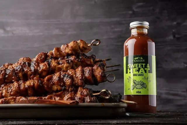 Grillede kjøttspyd stablet på en tallerken ved siden av en flaske Traeger Sweet & Heat BBQ Sauce 440 ml fra Traeger, alt mot en mørk bakgrunn.