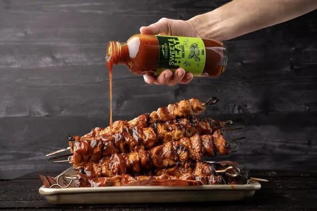 En hånd heller Traeger Sweet & Heat BBQ Sauce 440 ml over grillede kjøttspyd på en rektangulær tallerken, med den merkede sausen som gir en kraftig smak mot en mørk bakgrunn.