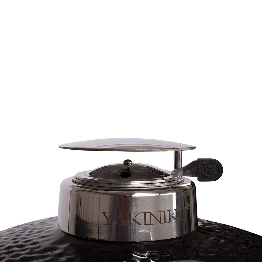 Nærbilde av en YAKINIKU Kamado Large 49 cm med en hel metallventilasjonshette på toppen av en svart keramisk grillplate med struktur.