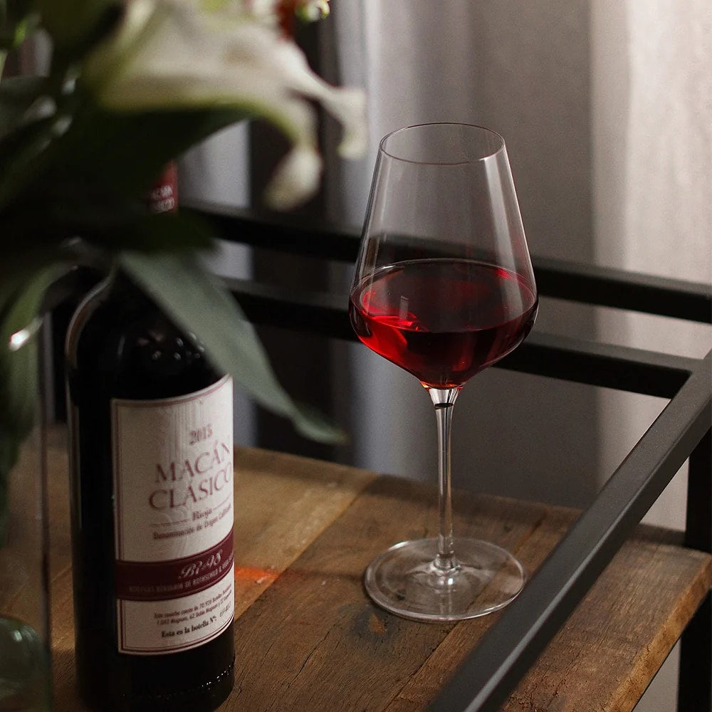 Et QUATROPHIL rødvinsvinglass 570 ml fra Stölzle Lausits, fylt med rødvin, står ved siden av en Macán Clásico-flaske på et trebord ved siden av en uskarp blomsterbukett.