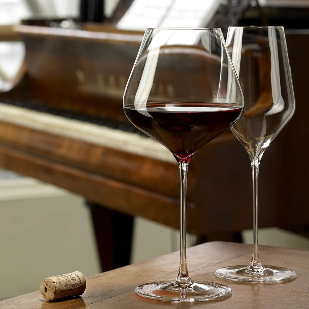 Et fylt QUATROPHIL Burgunder vinglass 710 ml fra Stölzle Lausits, et tomt, matchende glass og en kork står på et trebord foran et piano med noter.