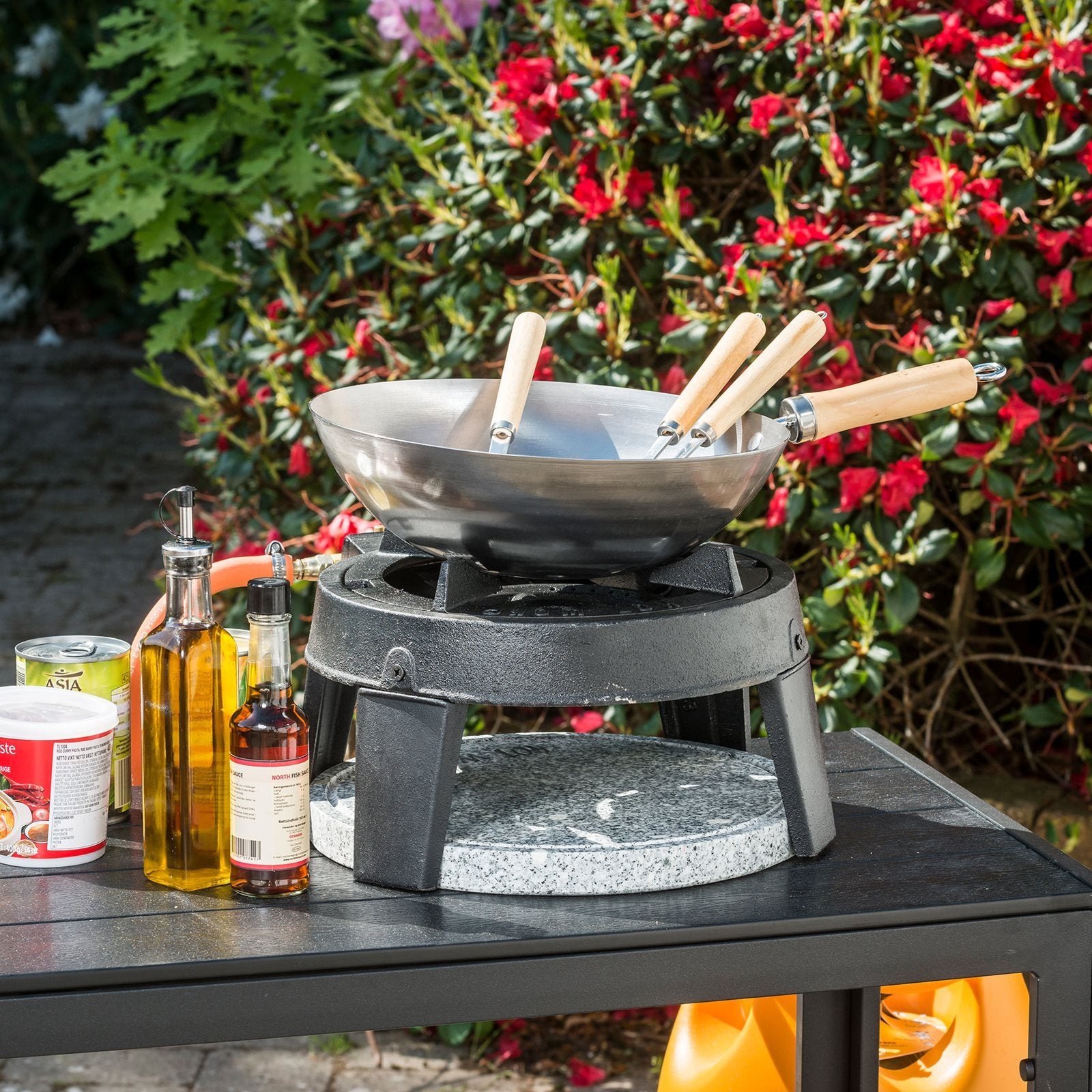A HOT WOK PRO 12 kW gassbrenner med wokpanne fra HotWok står utendørs omgitt av matoljer og hermetikk, med blomstrende busker i bakgrunnen.