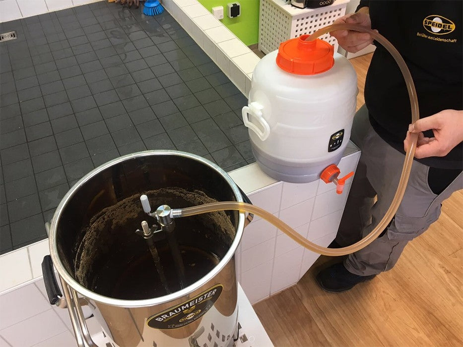 En person bruker Speidel Wort-pumpeenhet for 50L #Braumeister til å overføre væske gjennom et rustfritt rør til en Braumeister 50L bryggekjele i et flislagt rom.