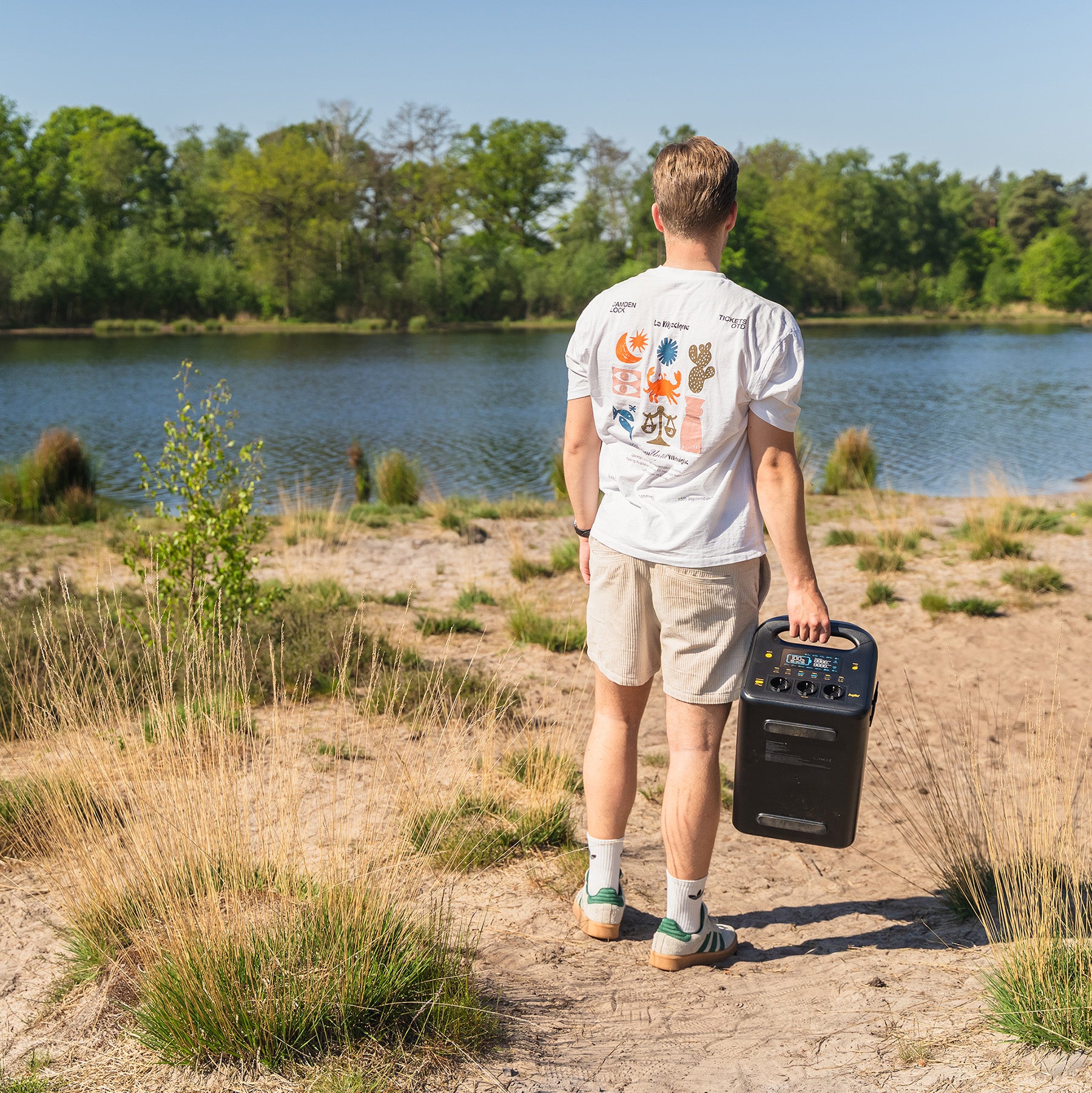 En person i hvit t-skjorte og shorts står på sandgrunn ved en innsjø med et Aqiila Powerbird S1800 v2 - Kraftstasjon nødbatteri fra Aqiila i hånden, med trær og gress i bakgrunnen.