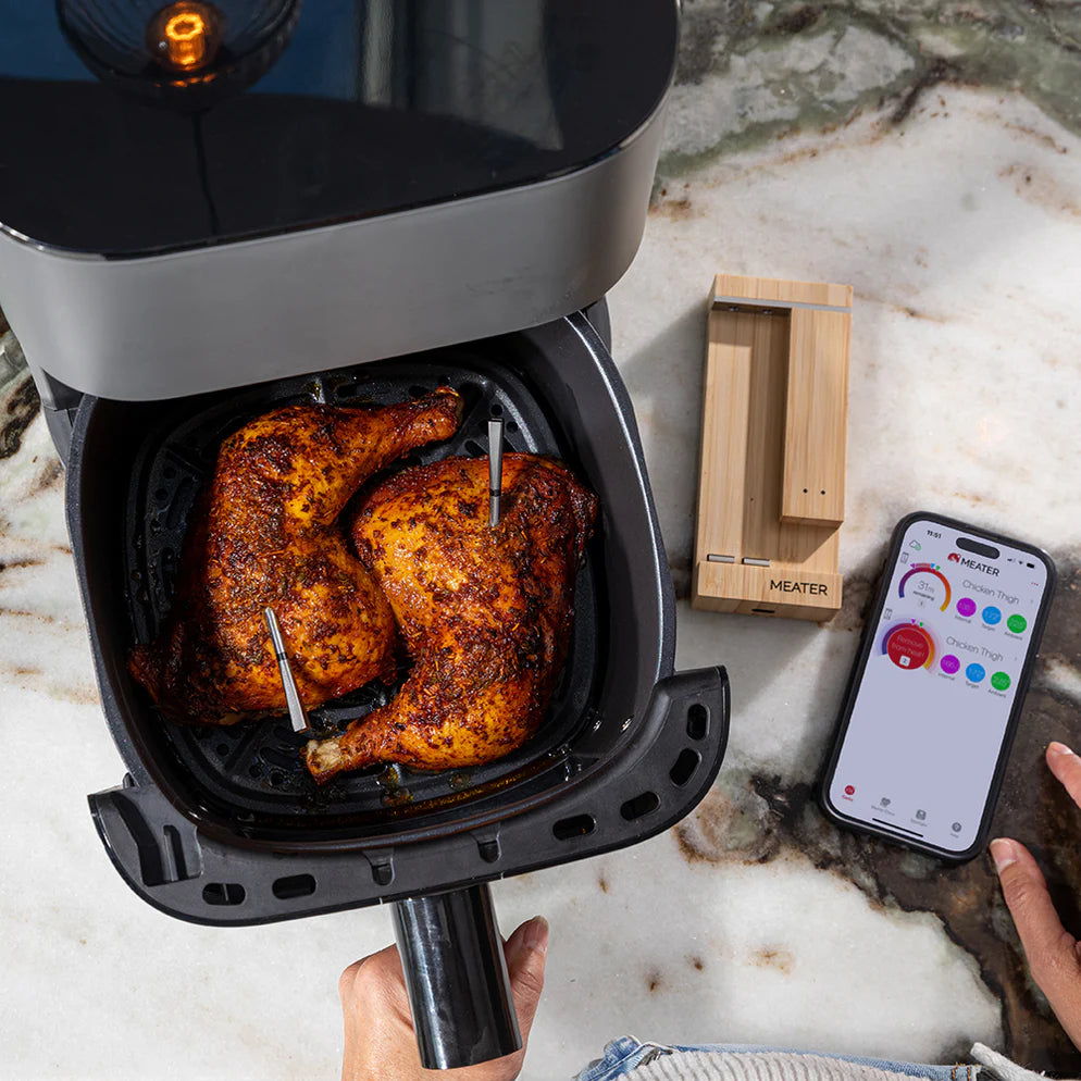 To stekte kyllinglår i en frityrgryte med kjøttermometer, en telefon som viser temperaturdata fra Traeger Meater Pro Duo - WiFi steketermometer, og den trådløse basen på en benkeplate i marmor.