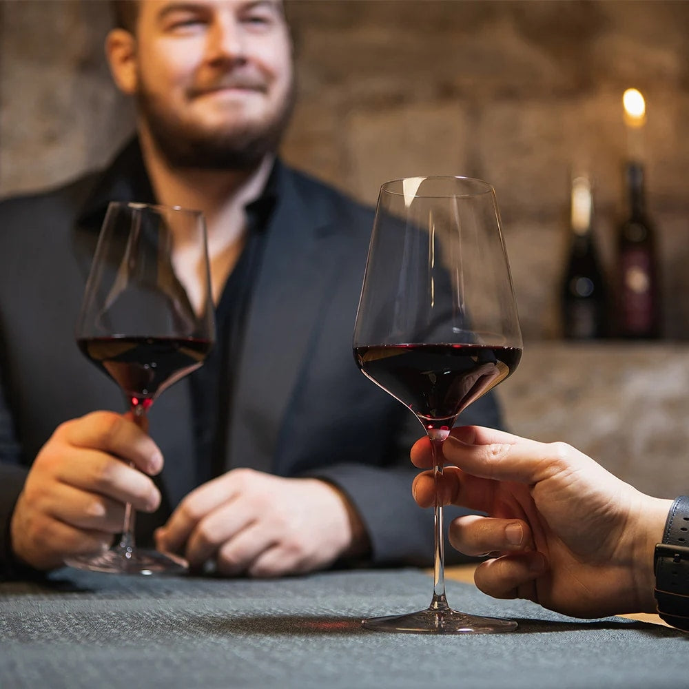 To personer sitter ved et bord med elegante STARlight Bordeaux vinglass 675 ml fra Stölzle Lausits, fylt med rødvin, mens vinflasker og et glødende stearinlys danner en varm, uskarp bakgrunn.