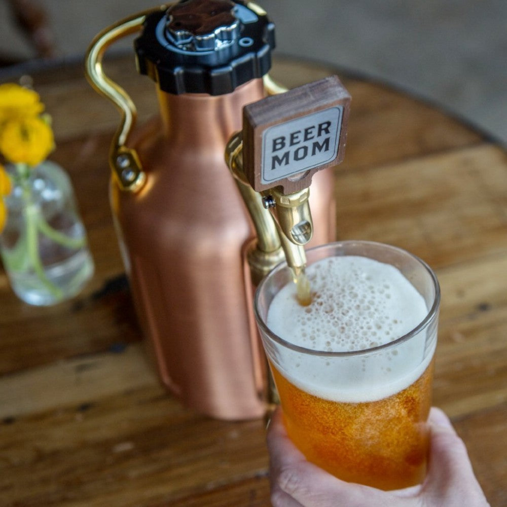 En hånd fyller et halvlitersglass med øl fra et GrowlerWerks uKeg utstyrt med Beer Mom Tap Handle på et trebord, ved siden av en skumbelagt eske.
