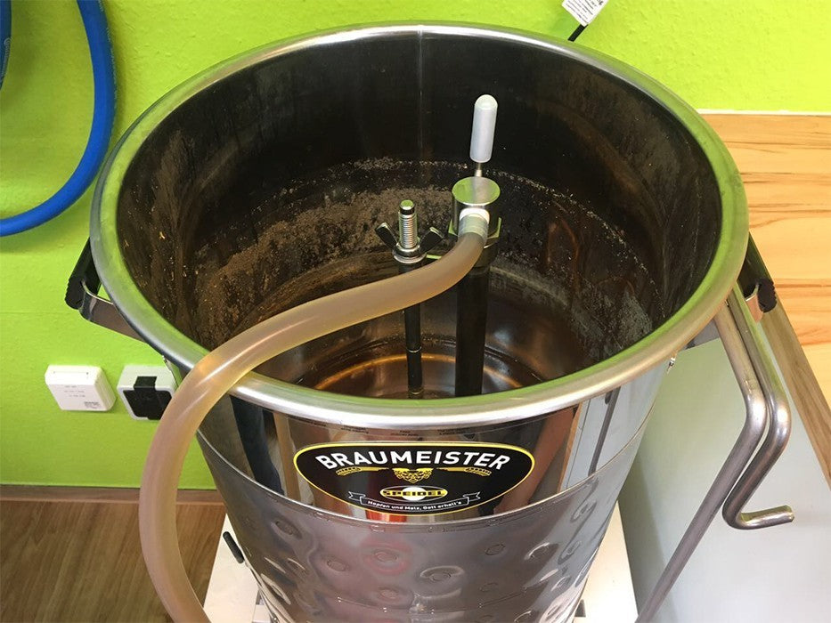 En Speidel Wort-pumpeenhet for 50L #Braumeister, komplett med slanger, koblinger og et rustfritt rør, står mot en grønn vegg i dette hjemmebryggingsoppsettet.