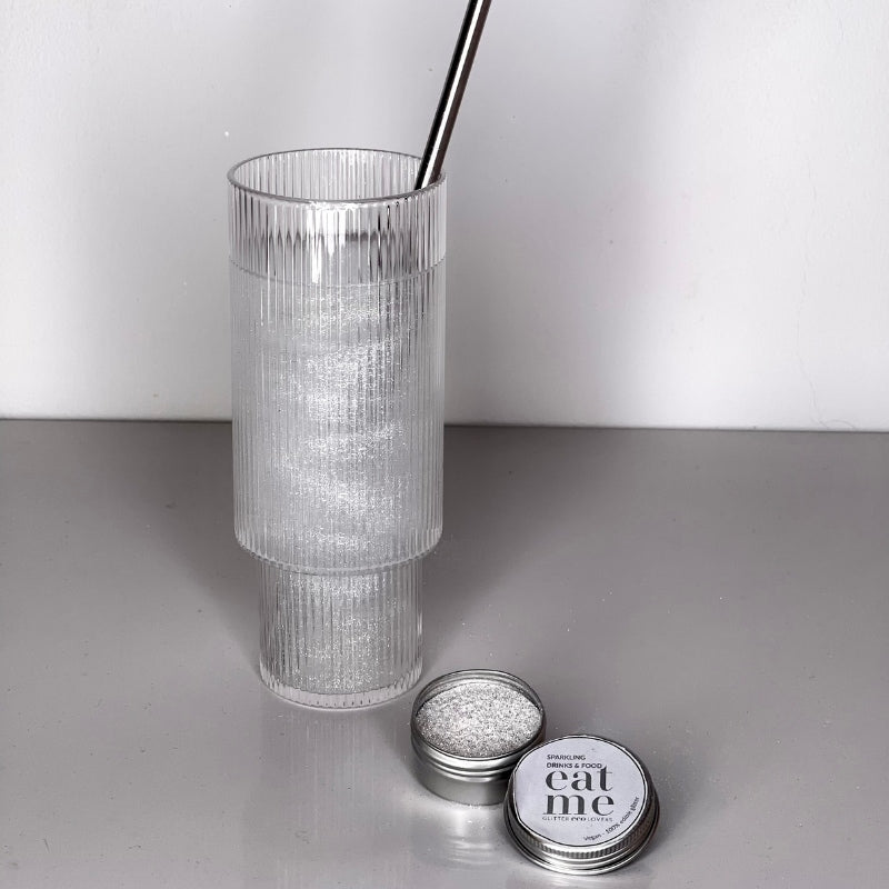Et klart glass med sugerør i metall inneholder en glitrende drink, ved siden av en åpen boks med Eco Lovers Eat Me Glitter Clear 20 ml fylt med spiselig sølvglitter - perfekt til å dekorere drinker eller kaker med.