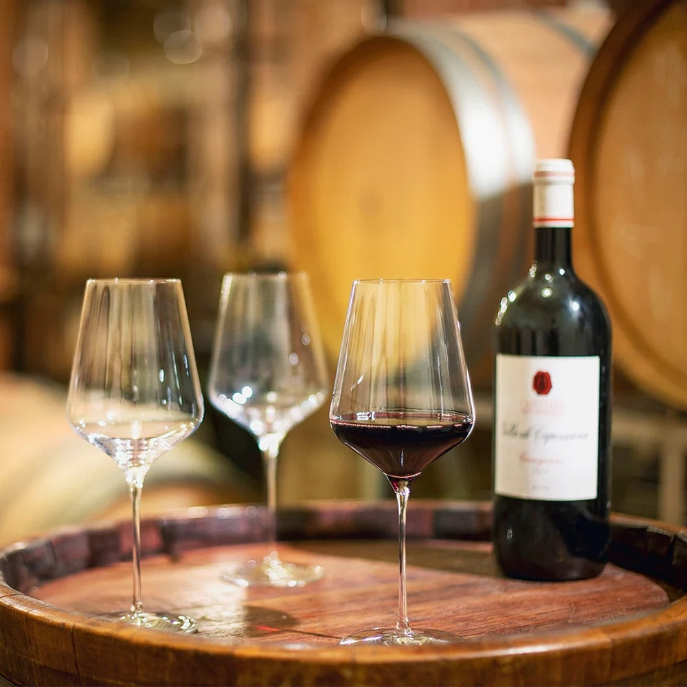 En vinflaske og tre Stölzle Lausitz STARlight Red Wine vinglass på 510 ml, ett fylt med rødvin, står på et trefat med uskarpe vintønner i bakgrunnen.