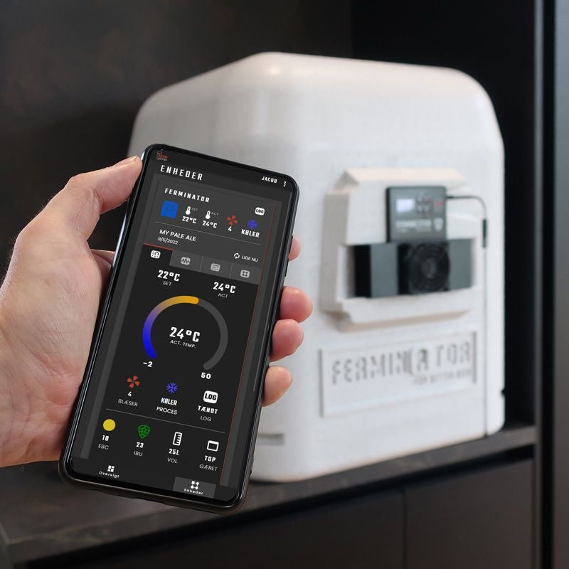 En person holder en smarttelefon som viser appen BrewCreator for gjæringskontroll med et Graviator WiFi-hydrometer og termometer fra Brewolution, foran en hvit gjæringskjele merket Ferminator.