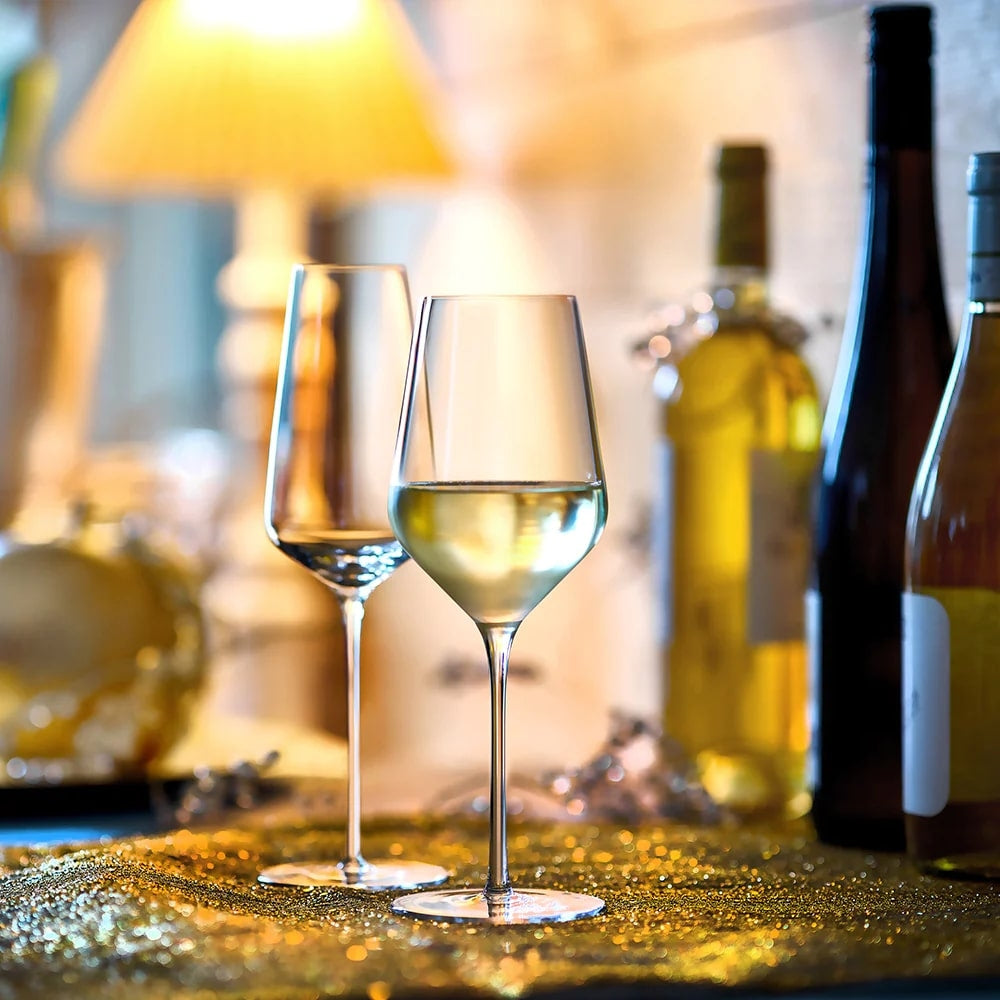 Et STARlight White Wine vinglass 410 ml fra Stölzle Lausitz, fylt med vin, står ved siden av et tomt glass på en glitrende gullflate, mens vinflasker og en svakt glødende lampe dukker opp i bakgrunnen.