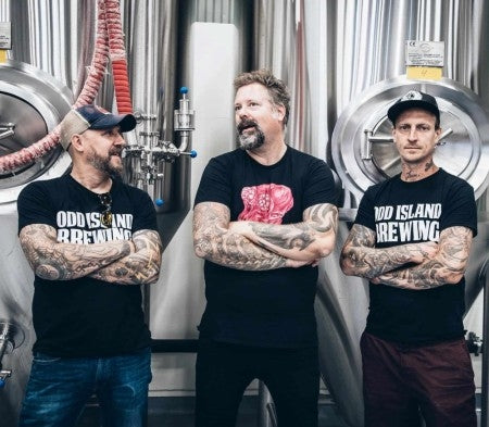 Tre tatoverte menn i Odd Island Brewing-skjorter står foran bryggetankene og viser frem sin håndverksinspirasjon - perfekt for fans av Odd Island Brewing Twelve Fifty - Allgrain ølsett eller andre vestkyst-IPA-entusiaster.