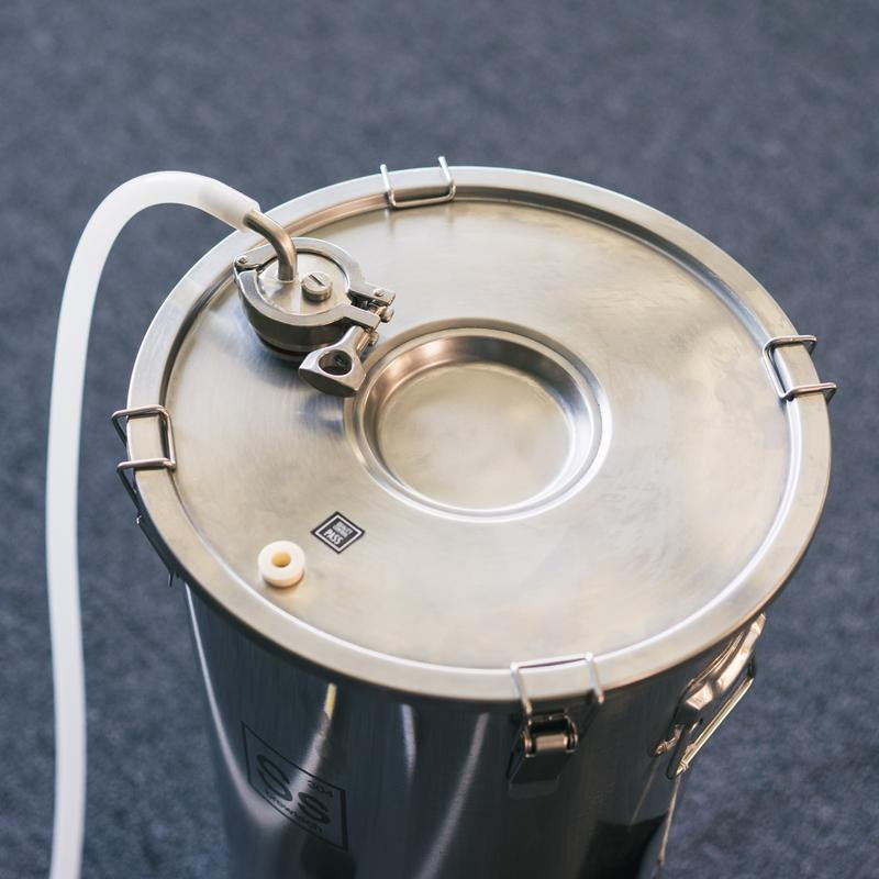En beholder av rustfritt stål 304 med forseglet lokk, slange festet til Brewshops 1,5" Tri-Clamp sveisefri gjennomføringsventil og flere låseklips, er plassert på et teppebelagt gulv.