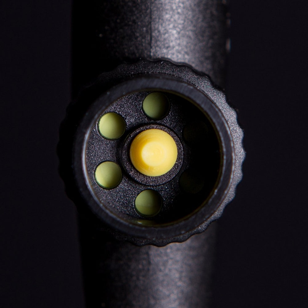 Nærbilde av en Plaato PLAATO Valve V2 - Pressure Drop Equalizing Valve, en svart, sirkulær enhet med seks grønne hull og en gul knapp i midten, vist mot en mørk bakgrunn.