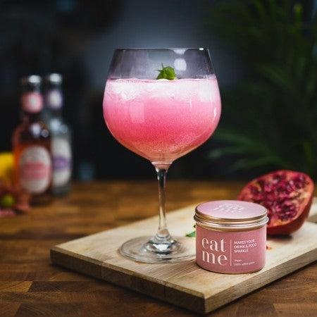 Et stort glass rosa drink med is, spiselig glitter og mynte står på en treplate ved siden av en "Eat Me Glitter Rose 20ml"-krukke fra Eco Lovers og et halvert granateple. I bakgrunnen skimtes uskarpe flasker.