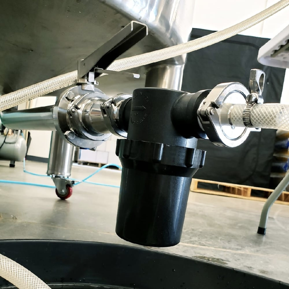 Nærbilde av et Bouncer Pro D Inline Filter 1,5" TC fra Bouncer festet til en tank i rustfritt stål med slanger koblet til på begge sider i et industrielt miljø for destillering av brennevin.