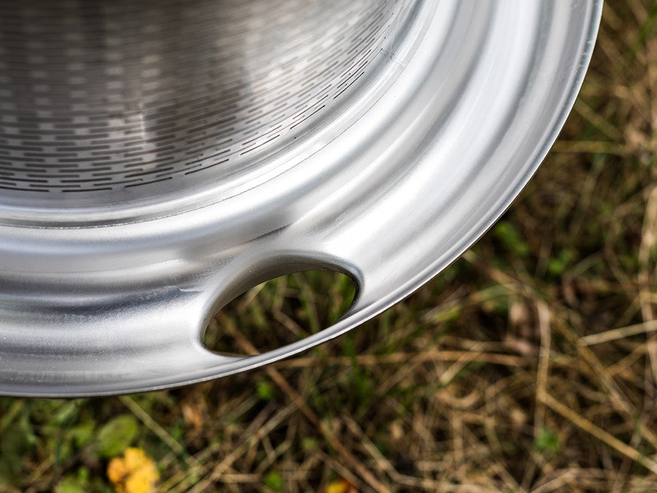 Nærbilde av en Speidel Hydropress 40L - Fruktpresse av rustfritt stål med perforert metalloverflate og stort hull nær kanten, plassert utendørs på tørt gress og spredt løv.