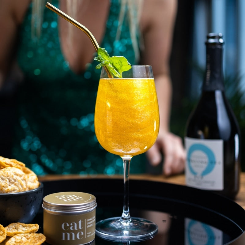 En person i grønn paljettkjole står bak en gyllen cocktail laget med Eco Lovers Eat Me Glitter Gold 150 ml, pyntet med mynte og servert ved siden av snacks, en lysboks og en vinflaske på et brett.