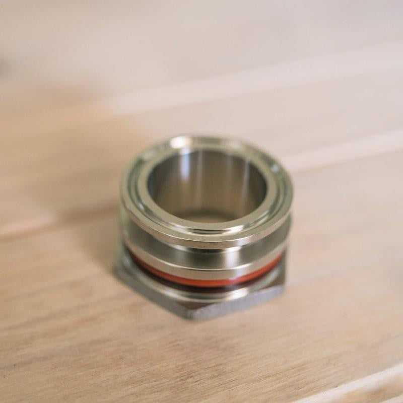 Brewshop 1,5" Tri-Clamp sveisefri gjennomføring, laget av 304 rustfritt stål med en sirkulær åpning og rød tetningsring, hviler på en lys treoverflate.