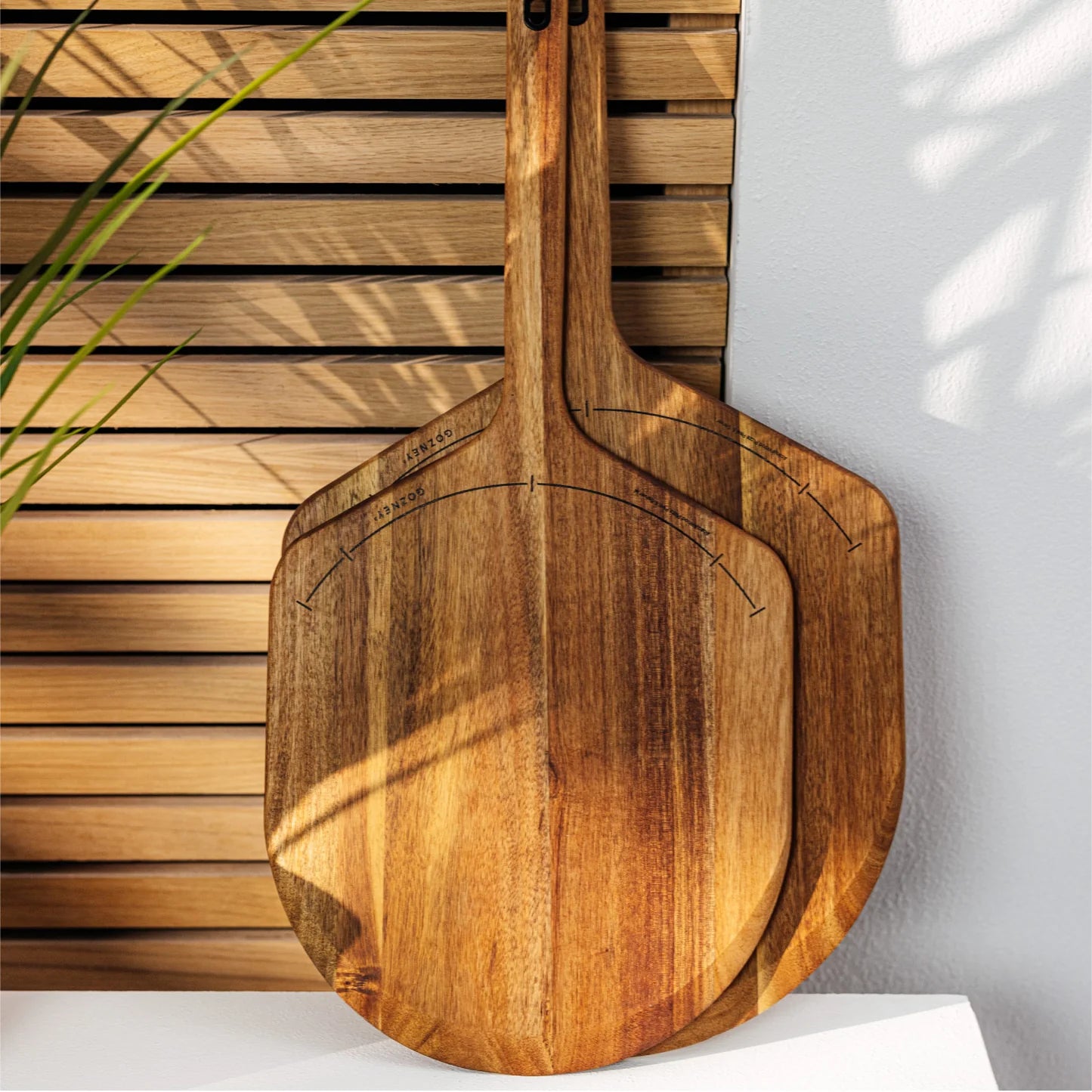 To Pizzaspade av tre, 14" Large (Pizzaspade av tre), laget av FSC-sertifisert akasietre, lener seg mot en spilevegg med sollys og en delvis synlig plante til venstre.