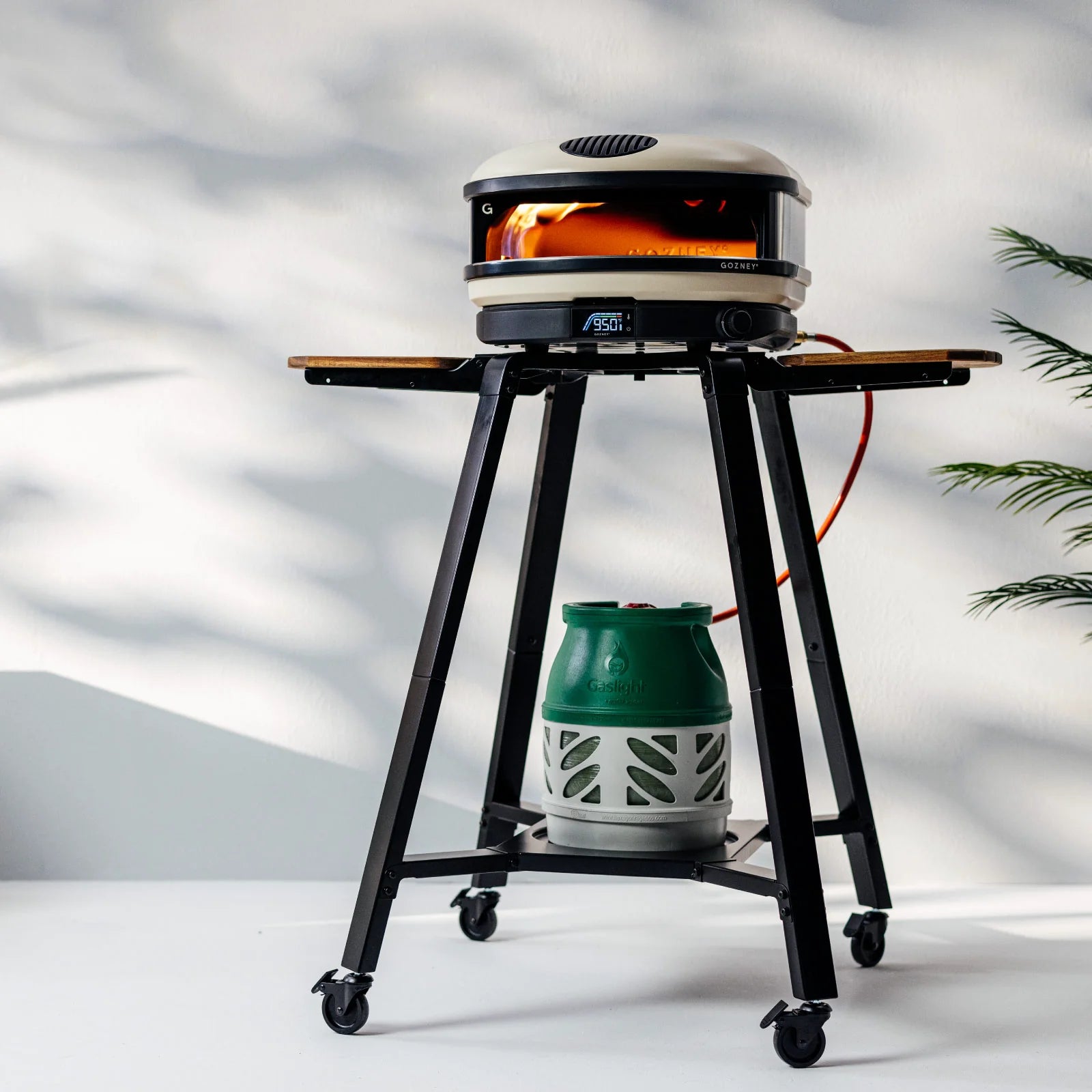 Pizzaovnen Gozney Arc står på toppen av Arc XL Stand, med en propantank under - perfekt for utendørs pizzabaking. En plante er synlig til høyre på en lys bakgrunn.
