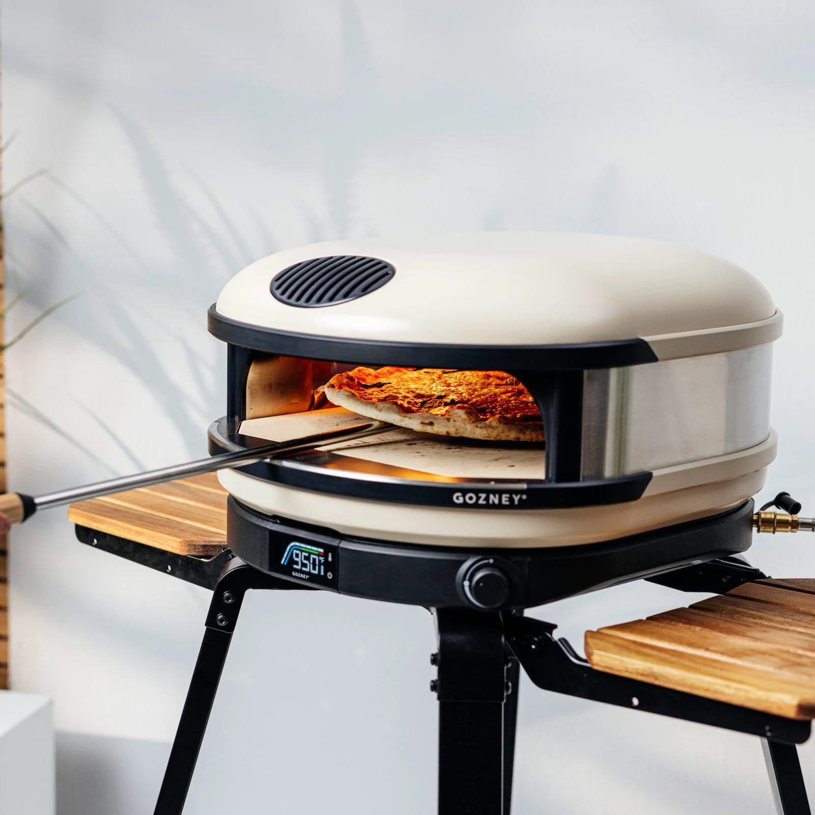 En pizza tas ut av en Gozney-pizzaovn på Gozney Arc + Arc XL Stand - Stativ til pizzaovn, med et digitalt display som viser 950°F-ideal for utendørs pizzabaking.
