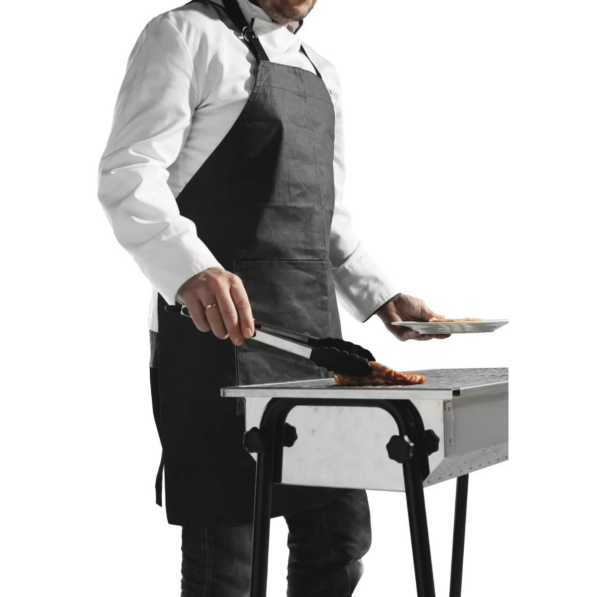 Iført Hendi BBQ Forkle og hvit skjorte bruker en person en grilltang til å grille mat, mens han holder en tallerken i den andre hånden.